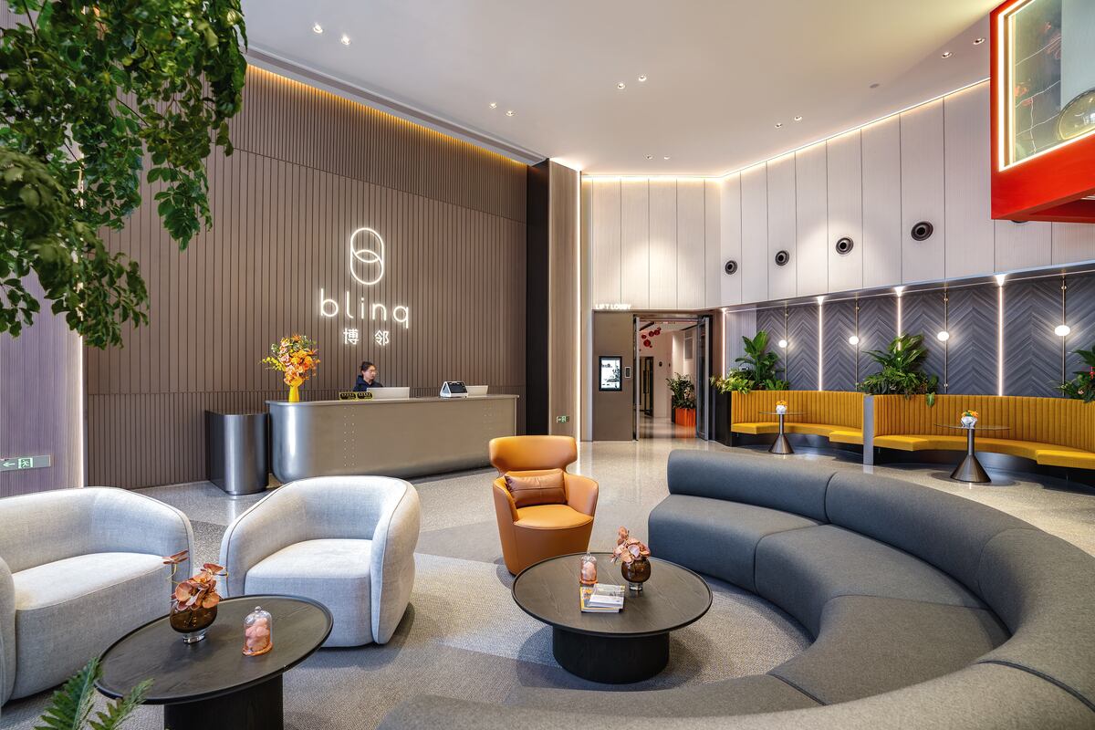 Stylish and modern office lobby featuring the "blinq" logo behind a reception desk. The space includes curved seating areas, contemporary furniture in neutral tones, warm lighting, potted plants, and a welcoming atmosphere designed for comfort and professionalism.