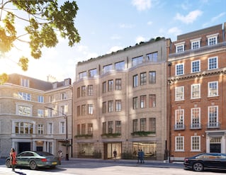 Photo of a light brown building with lots of windows and a couple cars in front.  Balcony at the top floor.  A light cream building on the left and a darker red brick building on the right