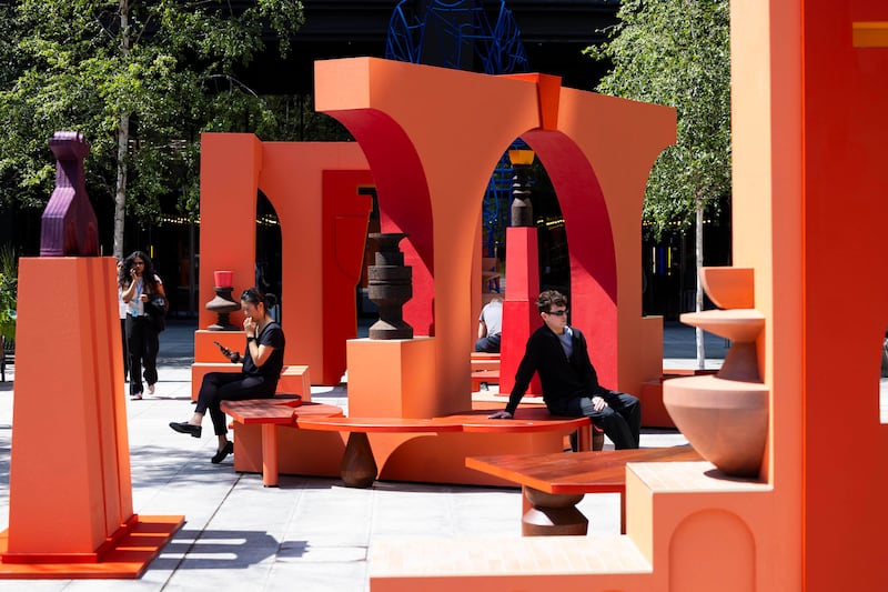 People sit and walk through a vibrant outdoor art installation composed of bold orange and red architectural forms, including arches, benches, and sculptural elements. The geometric structures create a playful, interactive space set against a backdrop of trees and urban surroundings.