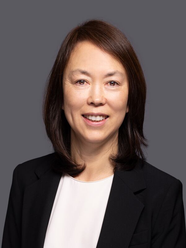 Professional headshot of a woman with straight, shoulder-length dark brown hair, wearing a black blazer over a white blouse. She is smiling and posed against a plain dark gray background.