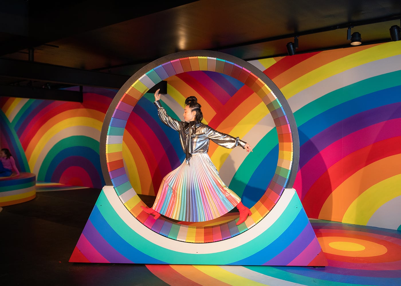 Rainbow Dreams by Hiromi Tango is art installation that is part of Luminato 2025. Pictured is the artist Hiromi Tango on a human size spinning hamster wheel. The rainbow wheel and the surrounding area is saturated with rainbow designs.