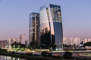 Two glass buildings tower over a river front. The sky is seen as a reflection in the side of one of the buildings.