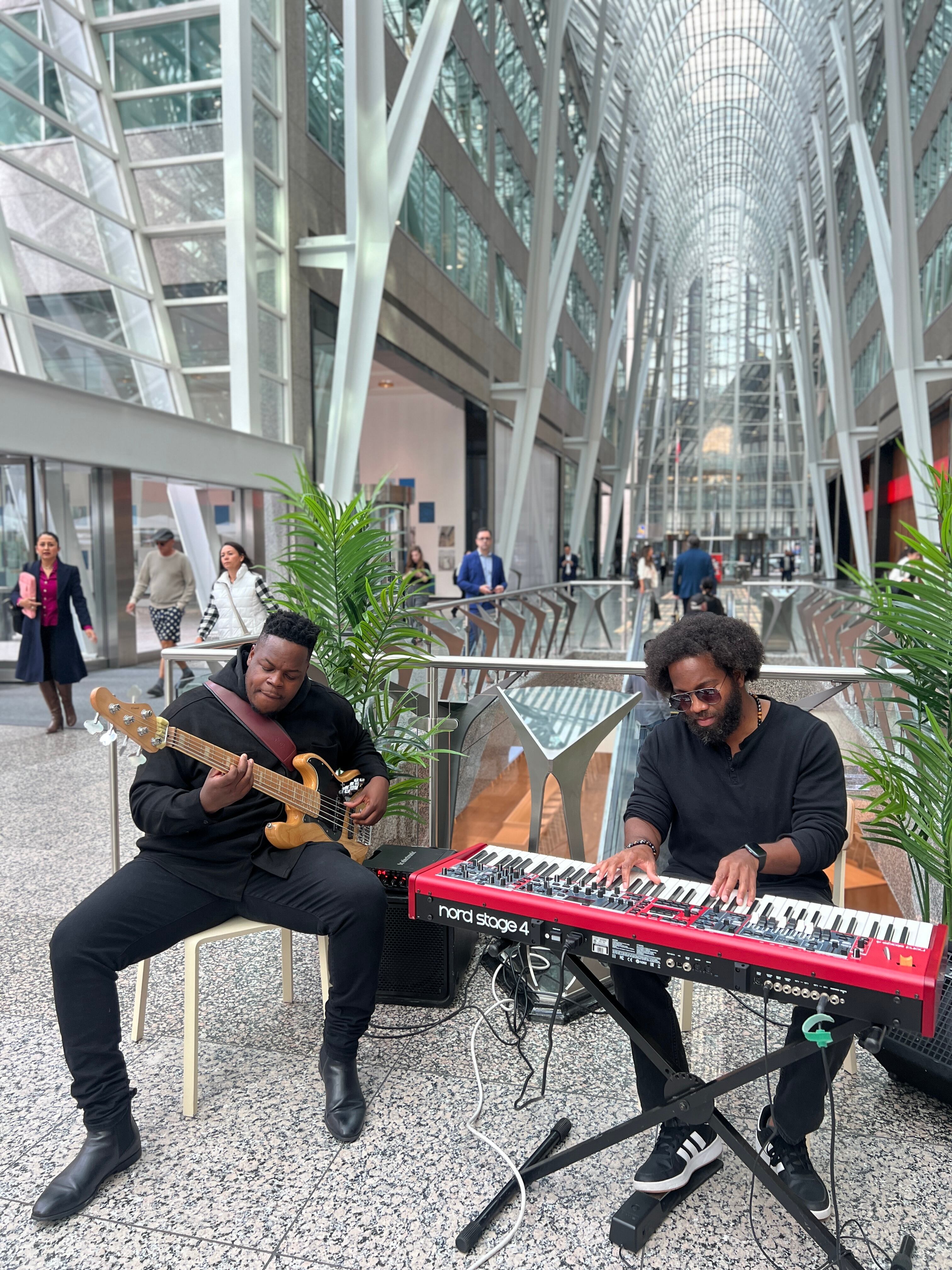 Two musicians, a guitar player and a pianist on an electro piano, in the middle of a busy day with lots of office workers walking through the Brookfield Place glass atrium.