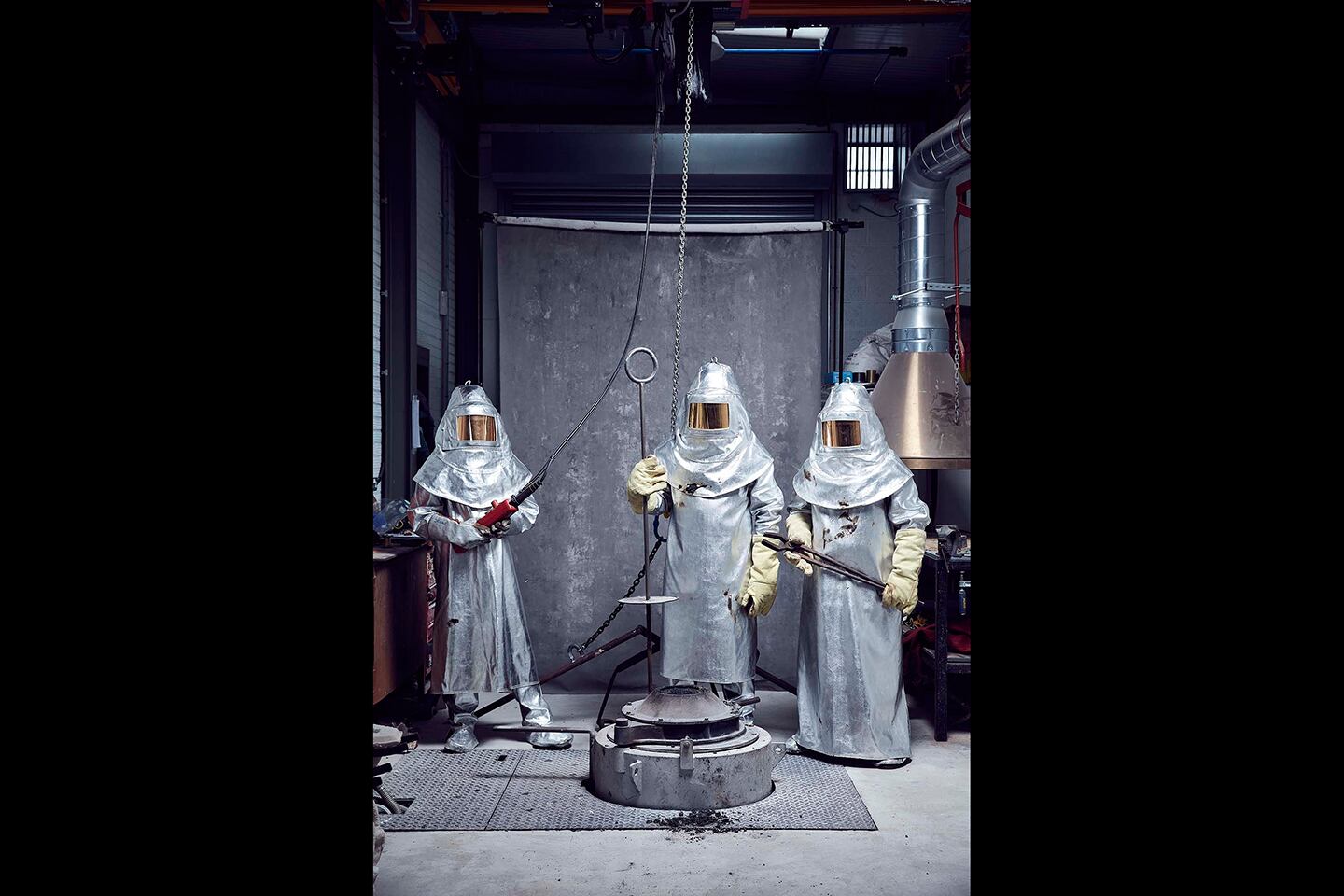 The image shows three people wearing full-body protective suits made of shiny, heat-resistant material with reflective visors and heavy gloves. They are standing in what appears to be an industrial or laboratory setting with concrete walls and metal equipment. The individuals are holding long metal tools, possibly used for handling extremely hot materials or working with molten metal. The equipment in front of them suggests a high-temperature process, likely involving casting or metalworking.