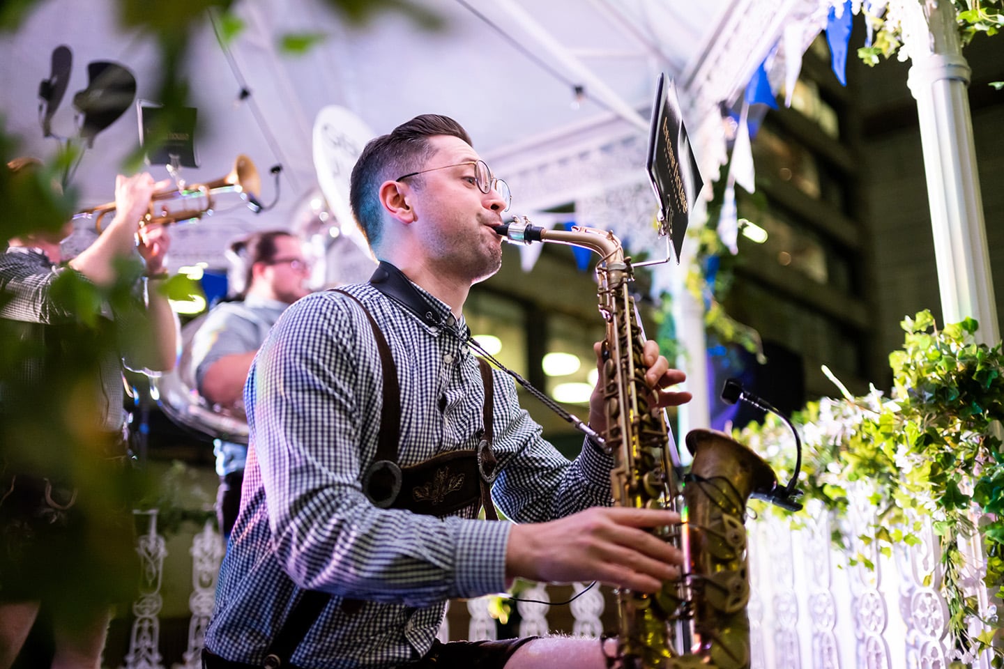A lively band performs at an outdoor Oktoberfest event. In the foreground, a musician wearing traditional Bavarian attire (checkered shirt with lederhosen and suspenders) plays the saxophone. Behind him, other band members play brass instruments. The stage is decorated with green ivy, blue-and-white Bavarian bunting, and ornate white railings. The atmosphere is festive and illuminated by night-time lighting.