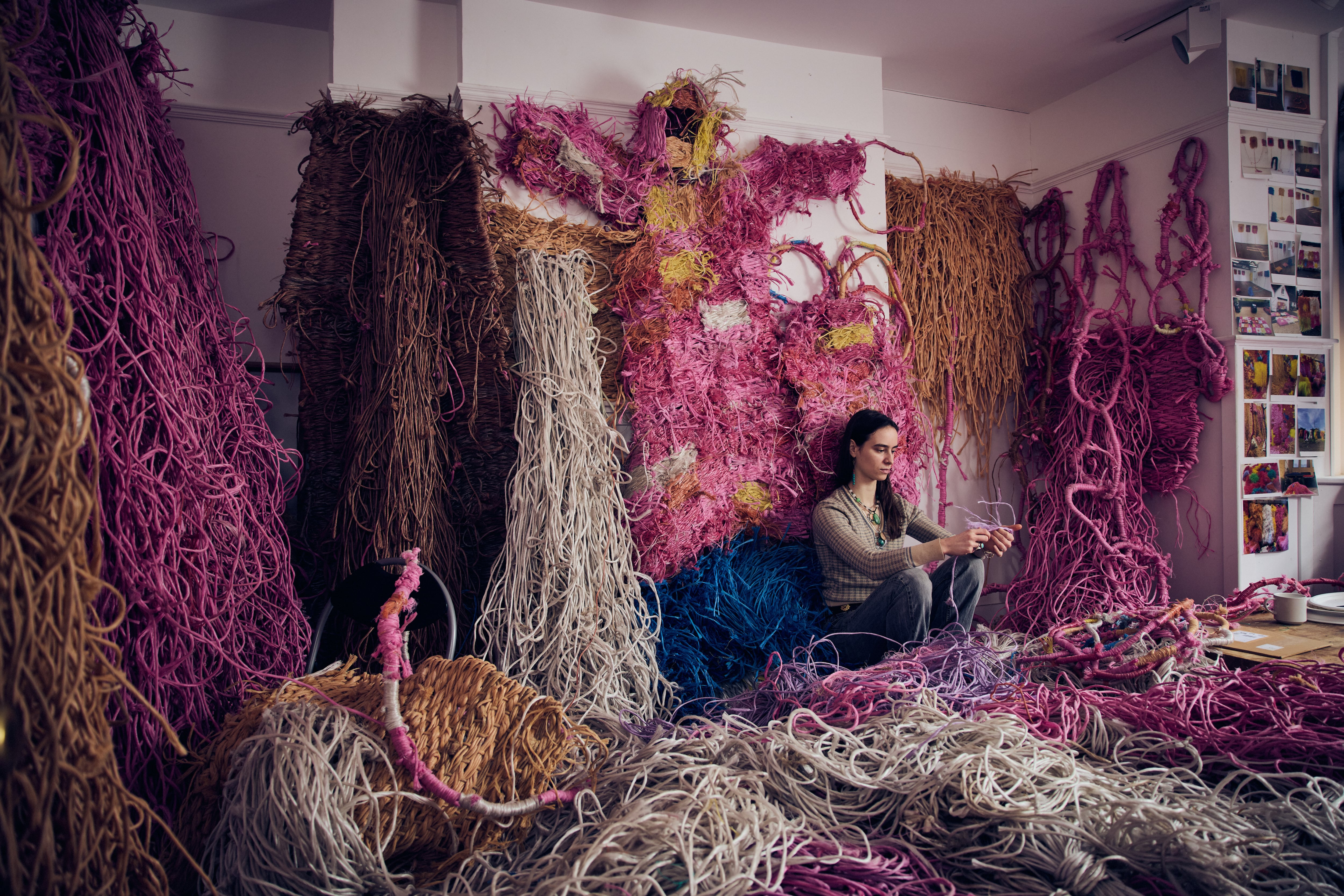Darcey Fleming sits cross-legged on the floor of a studio, surrounded by large, textured fiber artworks in shades of pink, brown, cream, and blue hanging on the walls. Piles of thick strands of rope or yarn cover the floor around her as she concentrates on weaving or tying a piece in her hands. Small photos and samples are pinned to the wall nearby, suggesting an active textile art workspace.