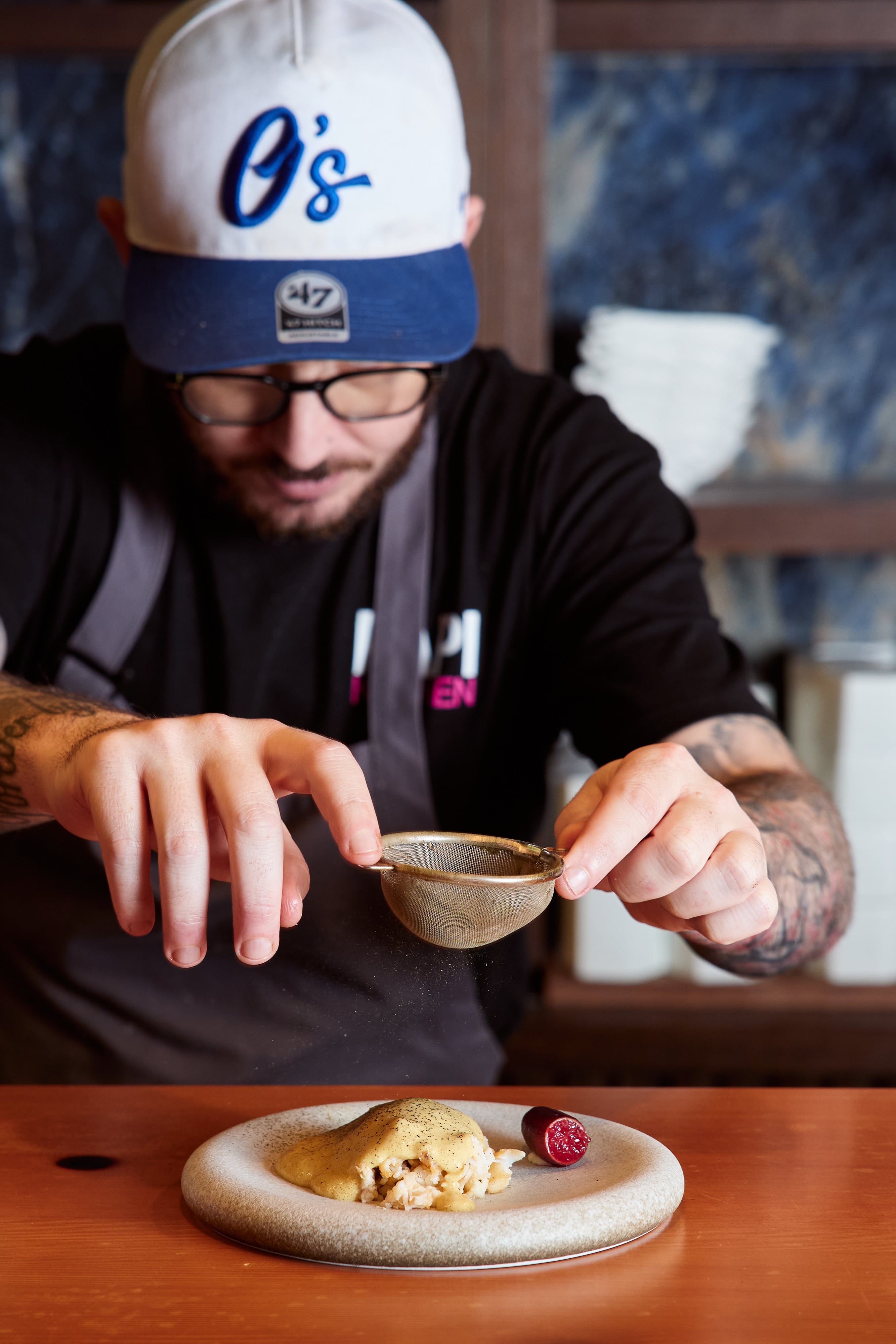 Man wearing a blue and white baseball cap holding a sifter over a plate of food.