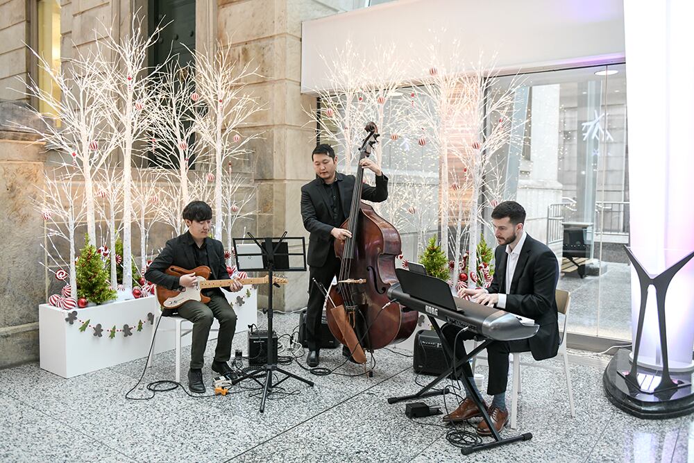Three musicians; a pianist, a basist and a guitarist play in front of some white, green and red holiday themed planters.