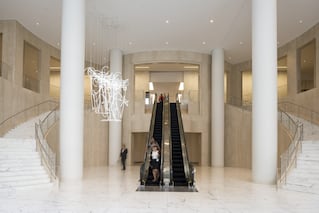 Interior of a building lobby with a set of marbled stairs on either side of an escalator directly in the center. A lighted sculpture hangs from the ceiling.