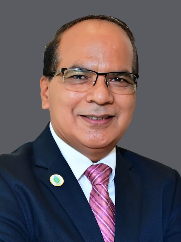 Man standing with a side pose.  Blue suit, white shirt, pink tie with a pin on his lapel