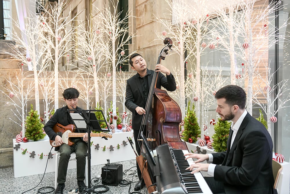 Three musicians; a pianist, a basist and a guitarist play in front of some white, green and red holiday themed planters.