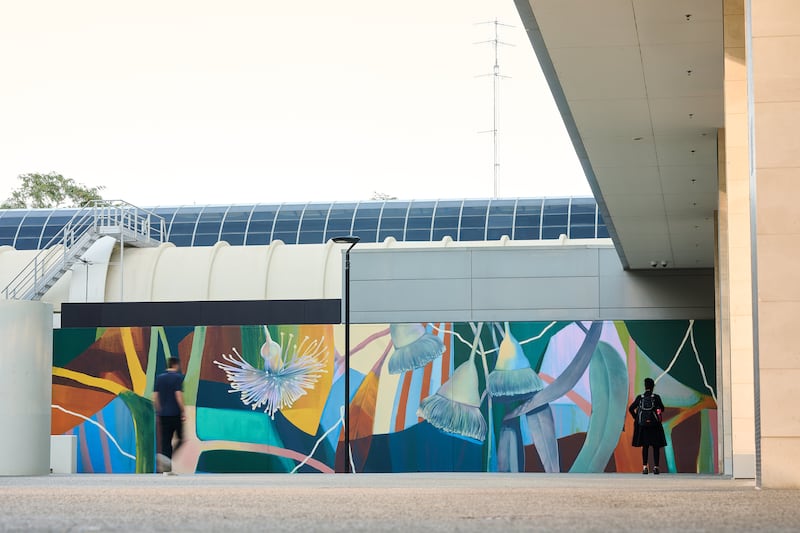 Wide view of a colorful botanical mural painted on an outdoor wall, featuring stylized native Australian flora with abstract leaves and large flower forms. The mural is situated in a modern urban setting with people walking by, and architectural elements like a curved roof and overhang frame the scene.