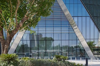 A large glass building that has an A shaped metal frame at the front of it and some trees out front as well.