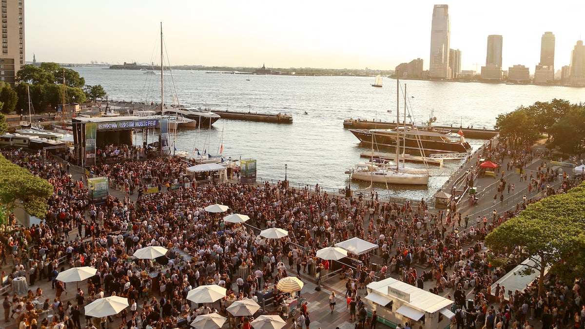 A crowd outside with water and boats in the background.  Buildings behind the water.