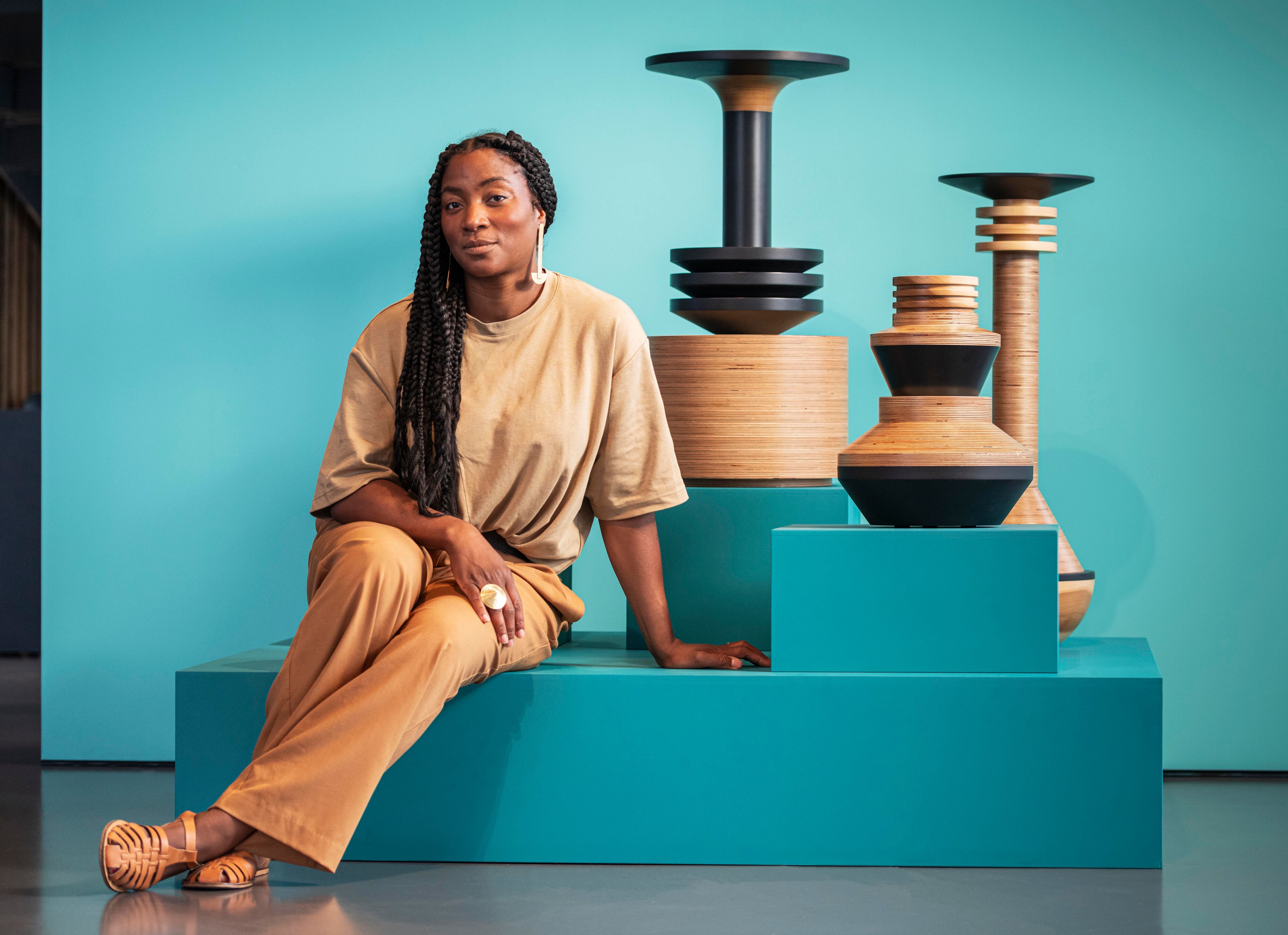 Turquoise background with 3 tan and black sculptures placed artistically. A woman with long black hair is sitting on a raised turquoise structure.