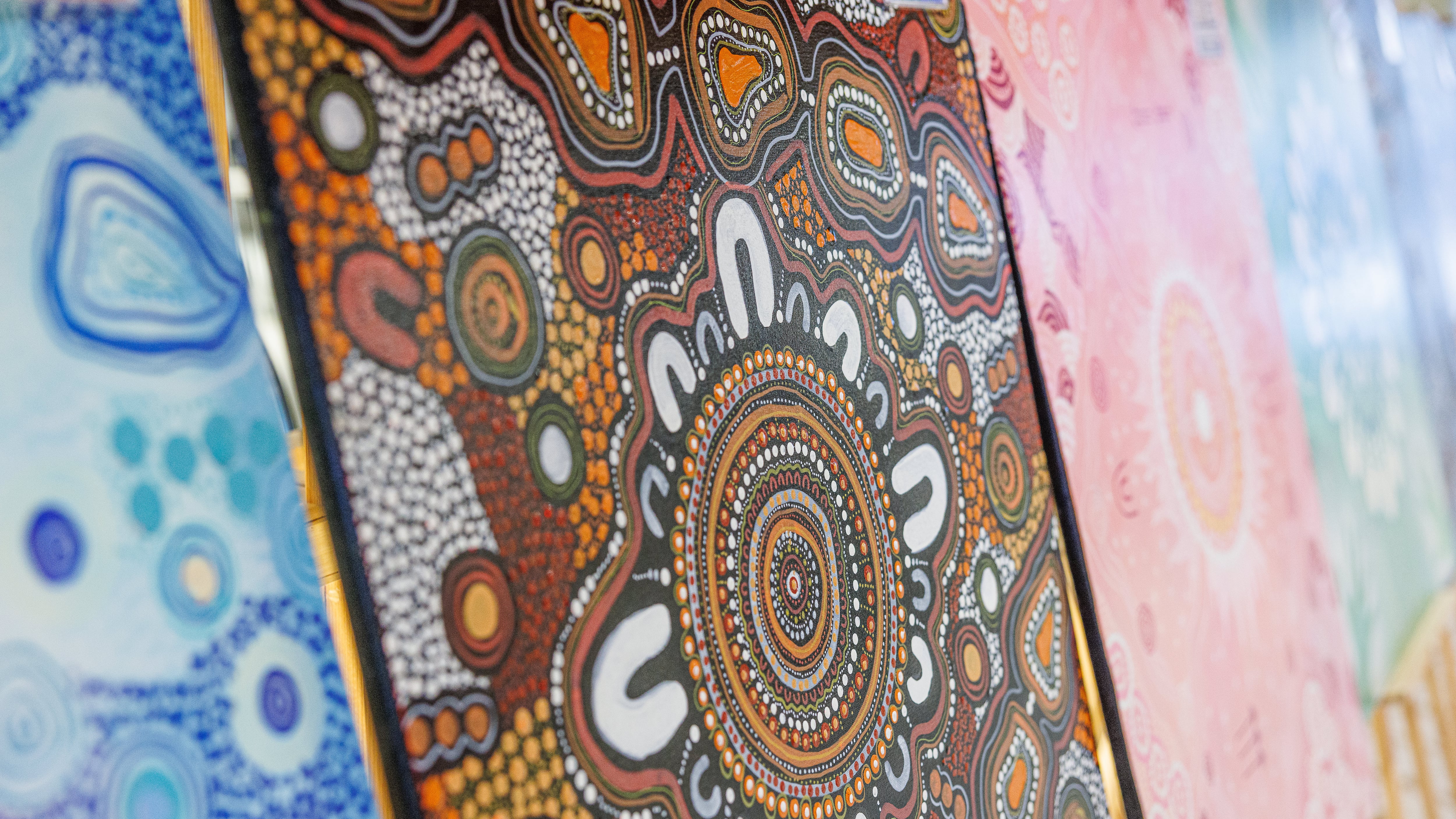 Close-up view of vibrant Aboriginal dot paintings displayed in a row, featuring intricate circular patterns and earthy tones. The central artwork includes detailed designs in brown, orange, white, and black, surrounded by other colorful paintings in blue, pink, and green hues.