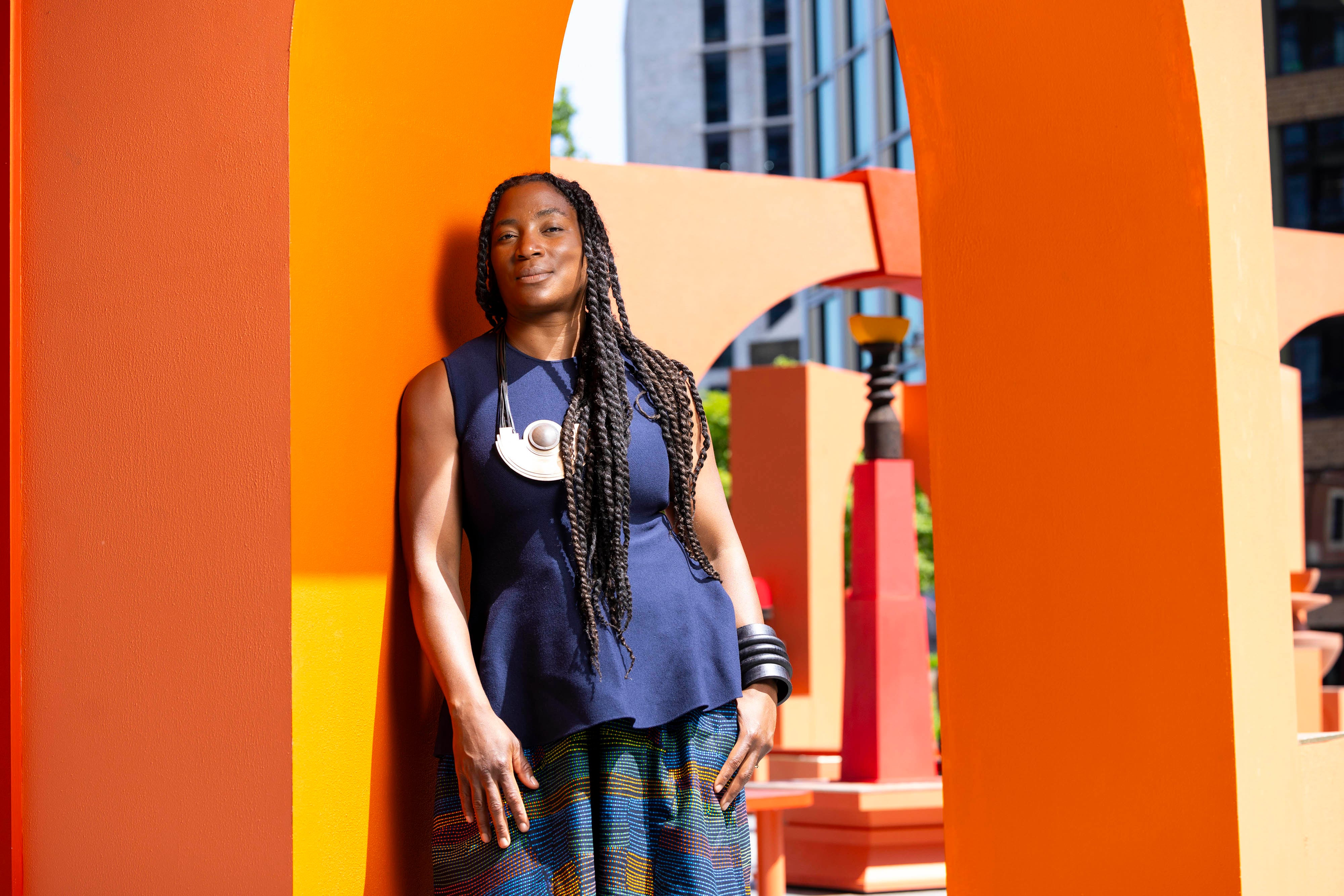 People sit and walk through a vibrant outdoor art installation composed of bold orange and red architectural forms, including arches, benches, and sculptural elements. The geometric structures create a playful, interactive space set against a backdrop of trees and urban surroundings.