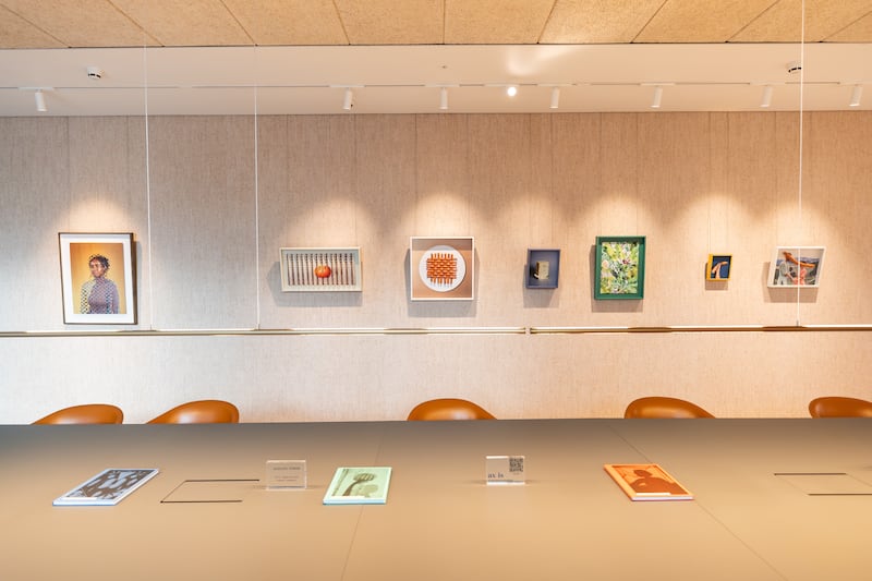A modern gallery-style conference room featuring a long table with books and placards, brown chairs, and a beige wall adorned with eight framed art pieces in various styles.