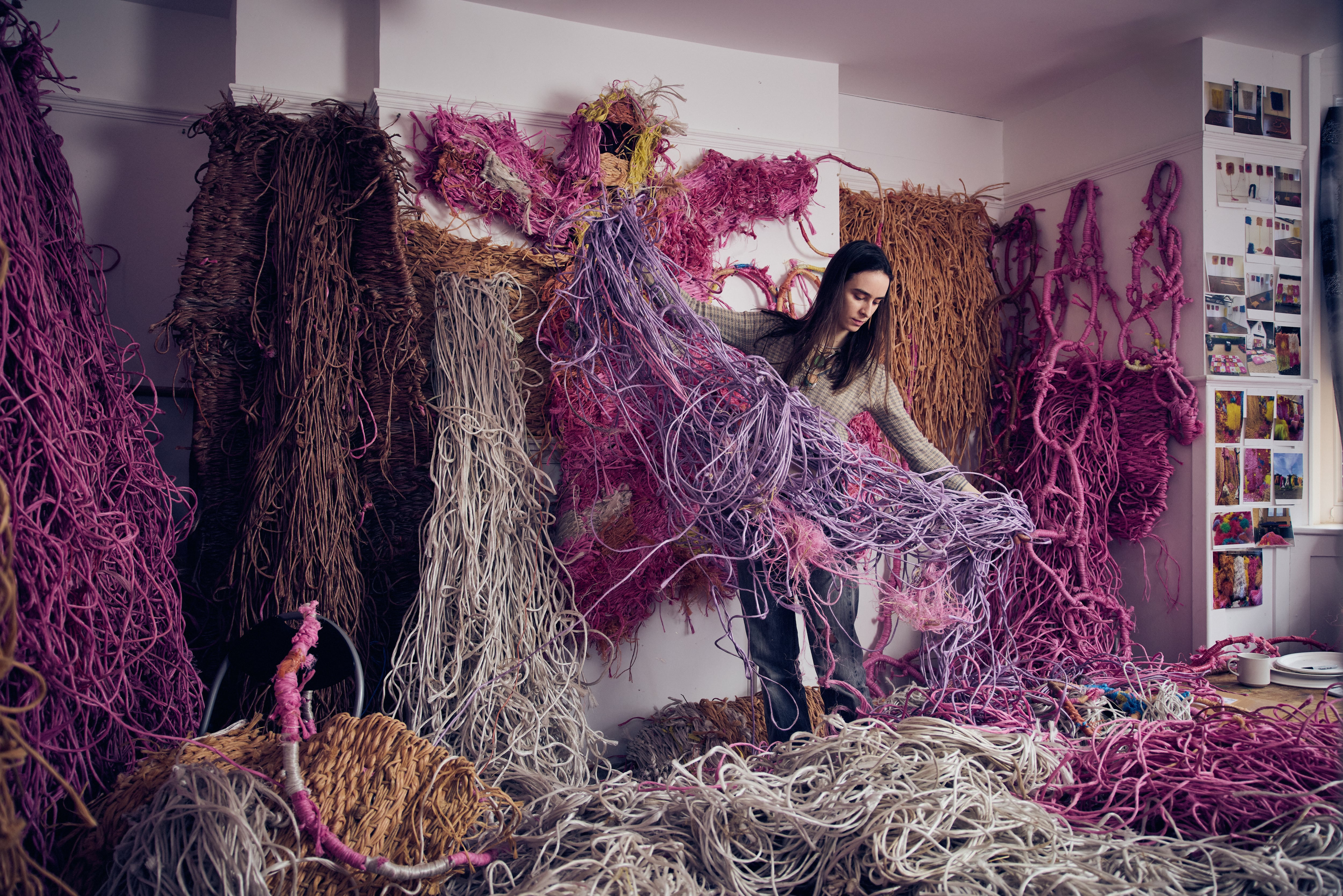 An artist stands in a studio surrounded by large, tangled textile installations in pink, brown, and cream tones, lifting a flowing mass of yarn-like fibres while similar woven pieces cover the walls and floor around her.