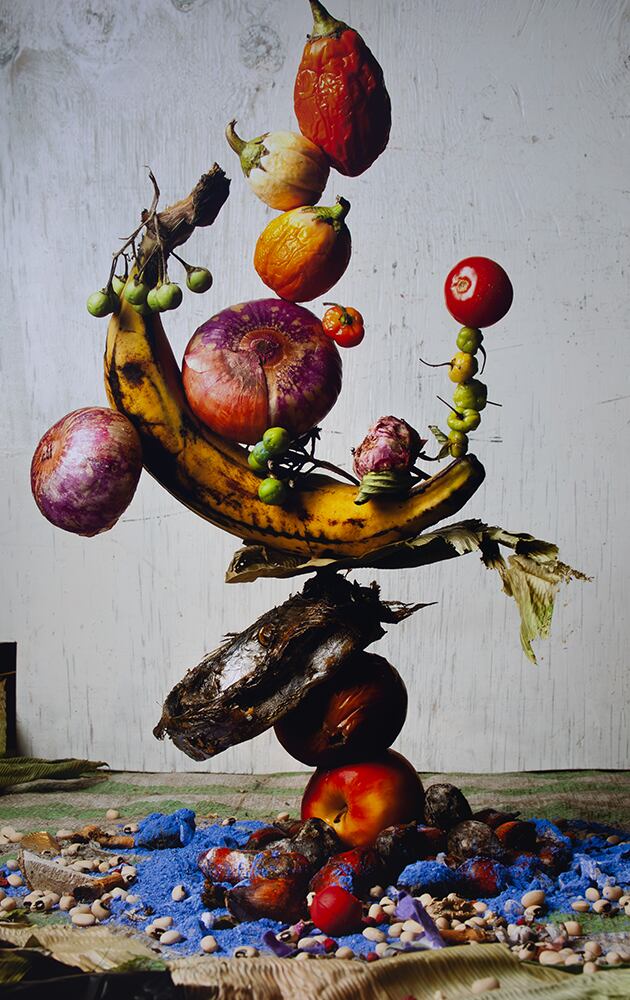 A visually striking sculpture made of balanced fruits and vegetables, including bananas, onions, tomatoes, and peppers, stacked vertically on a base of dried fish and apples. The arrangement rests on a textured mat sprinkled with blue pigment, beans, and organic materials, set against a plain white wall. The artwork merges natural decay with vibrant colour, suggesting themes of balance, impermanence, and renewal.