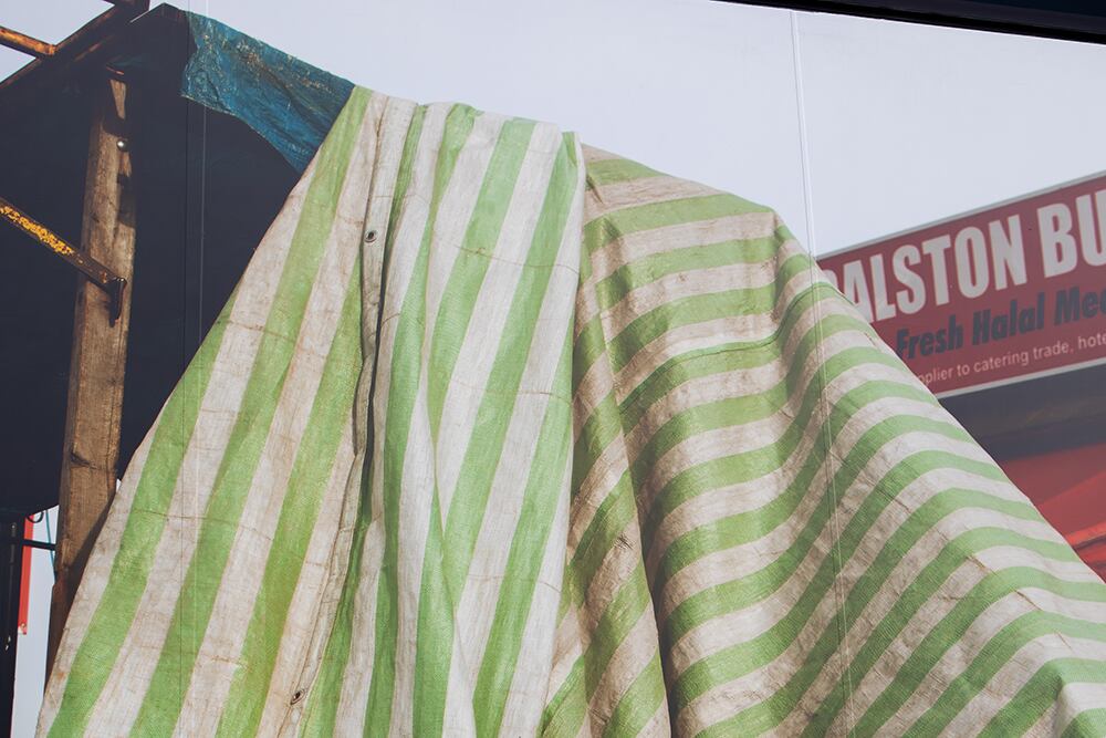 A large striped tarpaulin in green and white drapes over a wooden structure, partially covering a sign for “Dalston Butchers – Fresh Halal Meat.” The fabric appears weathered and sunlit, with folds and stains adding texture. The urban backdrop hints at a market or construction scene in an outdoor environment.