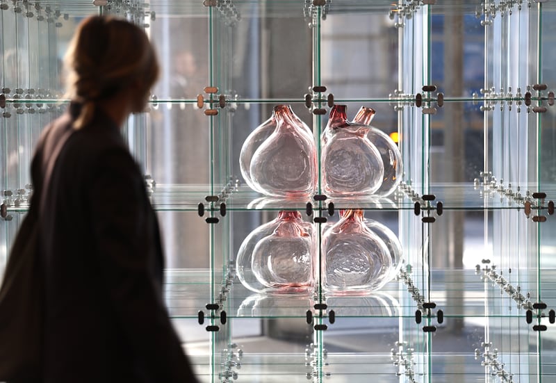 Woman blurred out in dark clothing standing in front of a shelf made up of square glass boxes carrying glass blown sculptures