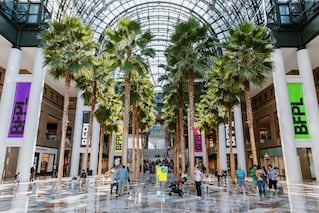 Trees that are in an indoor mall with people walking around on the tile flooring.