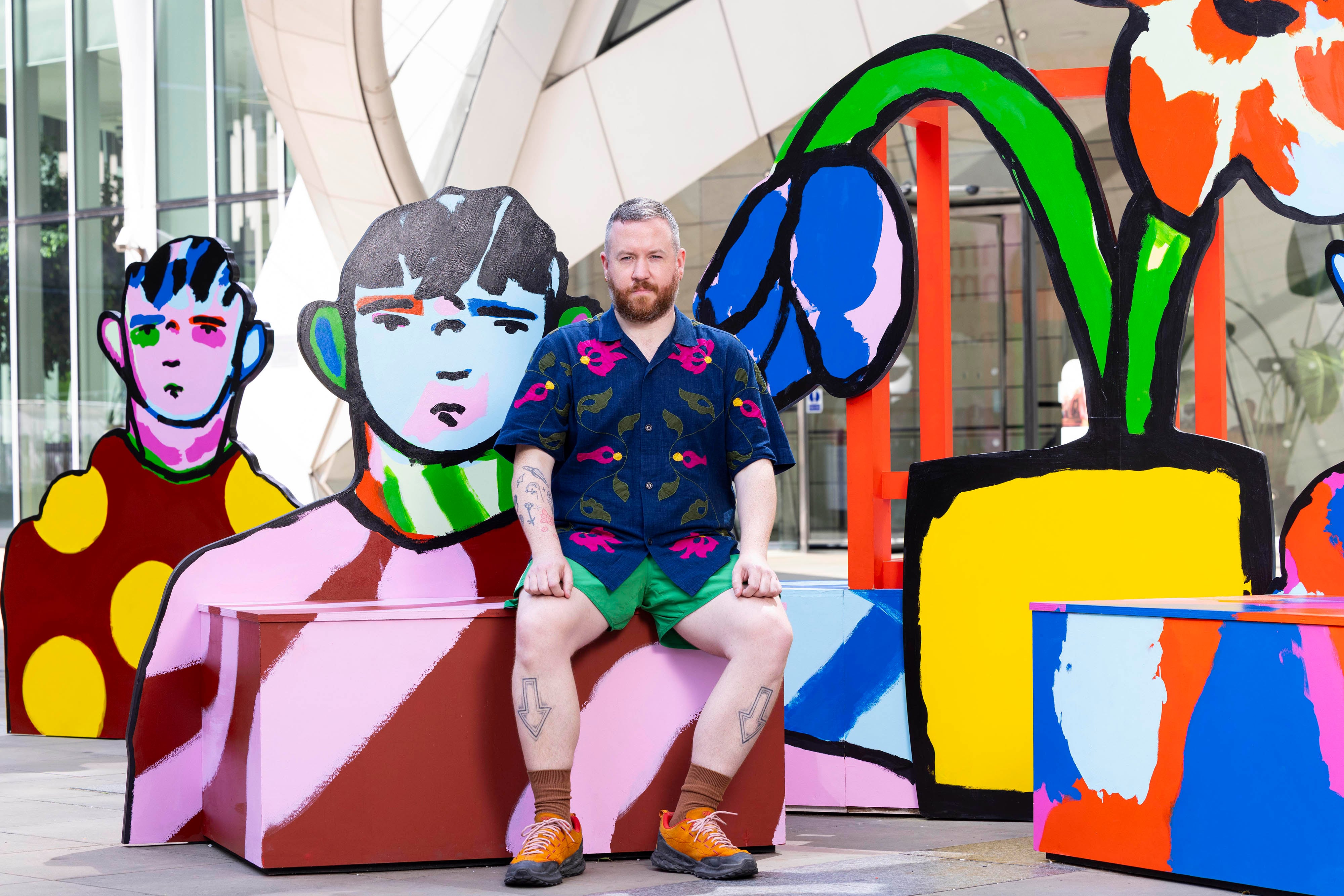 A man sits in front of a colorful public art installation featuring bold, cartoon-like figures and vibrant floral designs. The artwork uses thick black outlines and bright, saturated colors. The man is casually dressed in a patterned shirt and green shorts, blending with the playful, expressive aesthetic of the scene.