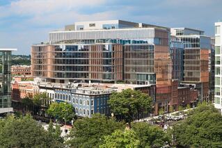 A multilayered building with many windows sits in the city. It has many trees around it.