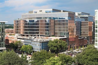 A multilayered building with many windows sits in the city. It has many trees around it.