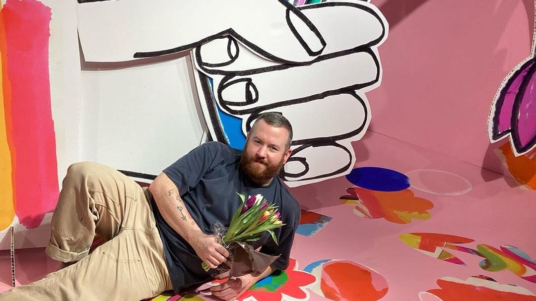 A large drawing/art installment, a large hand that is holding a selection of colorful flowers against a pink background. A gentleman on the floor holding a bouquet of flowers.
