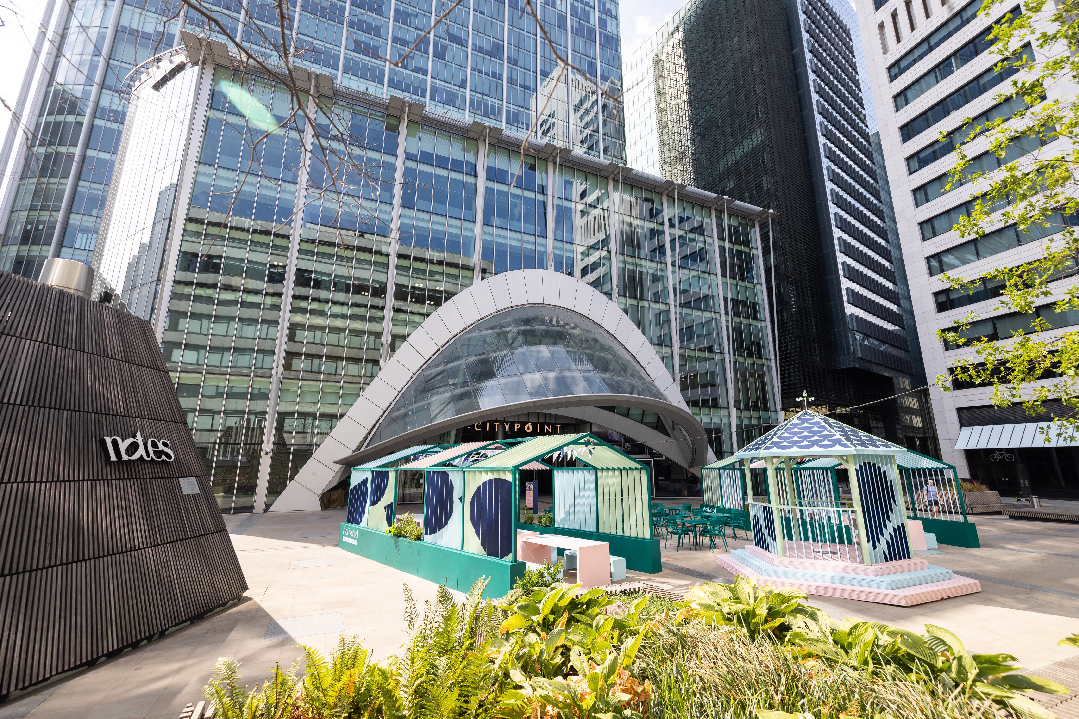 A modern urban plaza surrounded by tall glass-and-steel office towers. At the center is a large arched glass entrance labeled “CITYPOINT,” set into a multi-story office building. In front of the entrance are two decorative pavilion-style structures painted in pastel turquoise, navy, and white geometric patterns, with outdoor café-style seating between them. On the left stands a dark, sculptural cone-shaped feature displaying the word “notes.” Low green plants and ferns fill the foreground, while tree branches frame the top edges of the image. The scene is brightly lit by sunlight, creating reflections on the surrounding skyscraper façades.