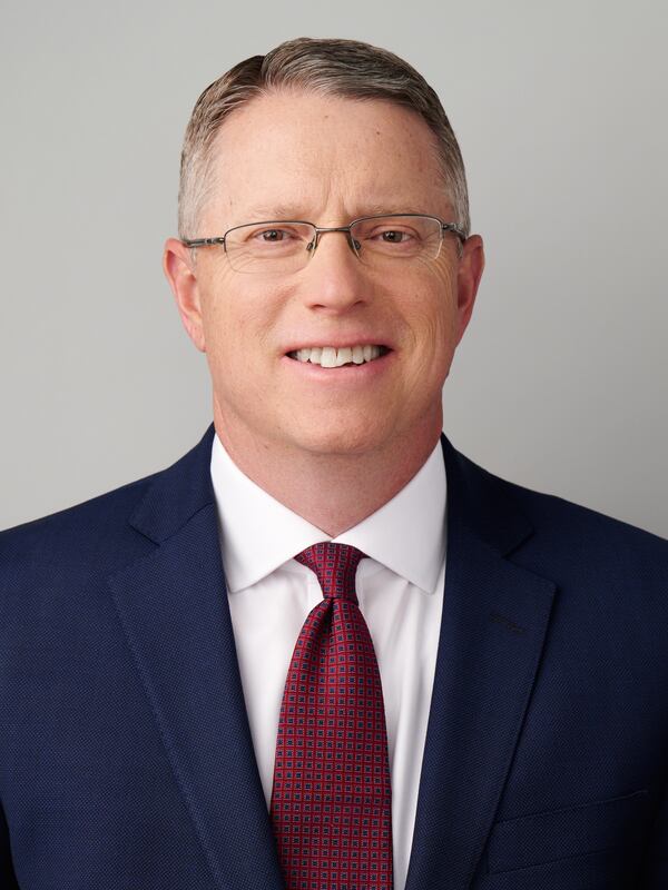 A white man with a blue blazer, white shirt and maroon tie wearing glasses