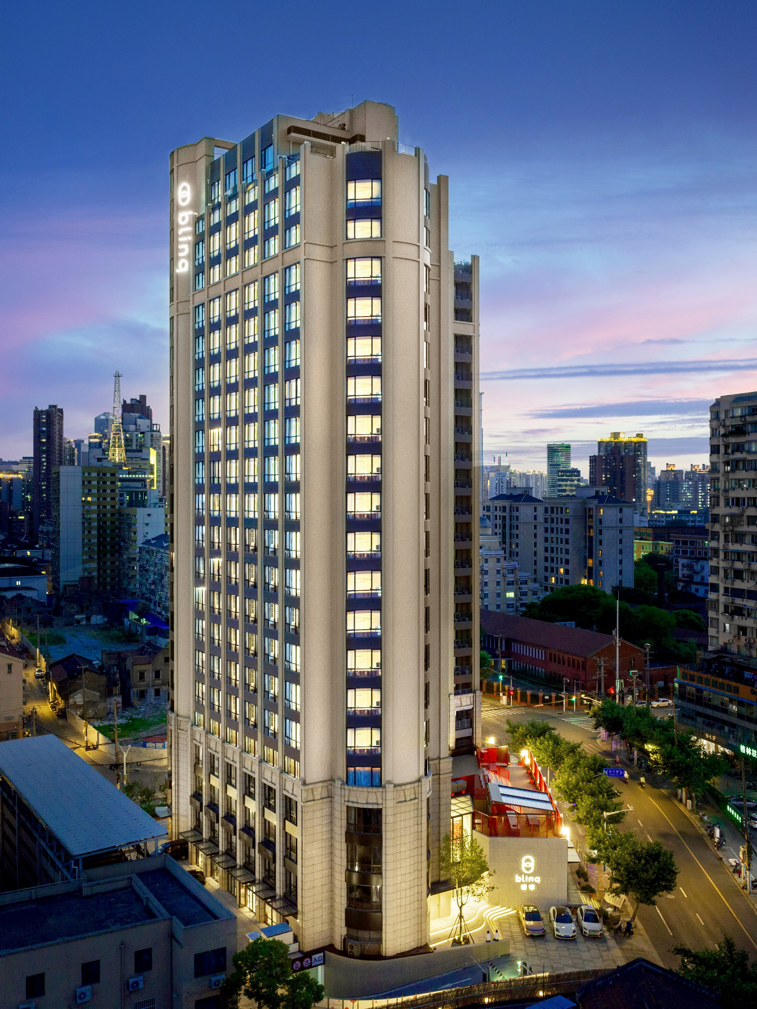 Nighttime view of a tall, modern high-rise building with illuminated windows, situated at a busy street intersection in an urban cityscape. The sky is a blend of blue and pink hues, and surrounding buildings and streets are lit with ambient lighting.