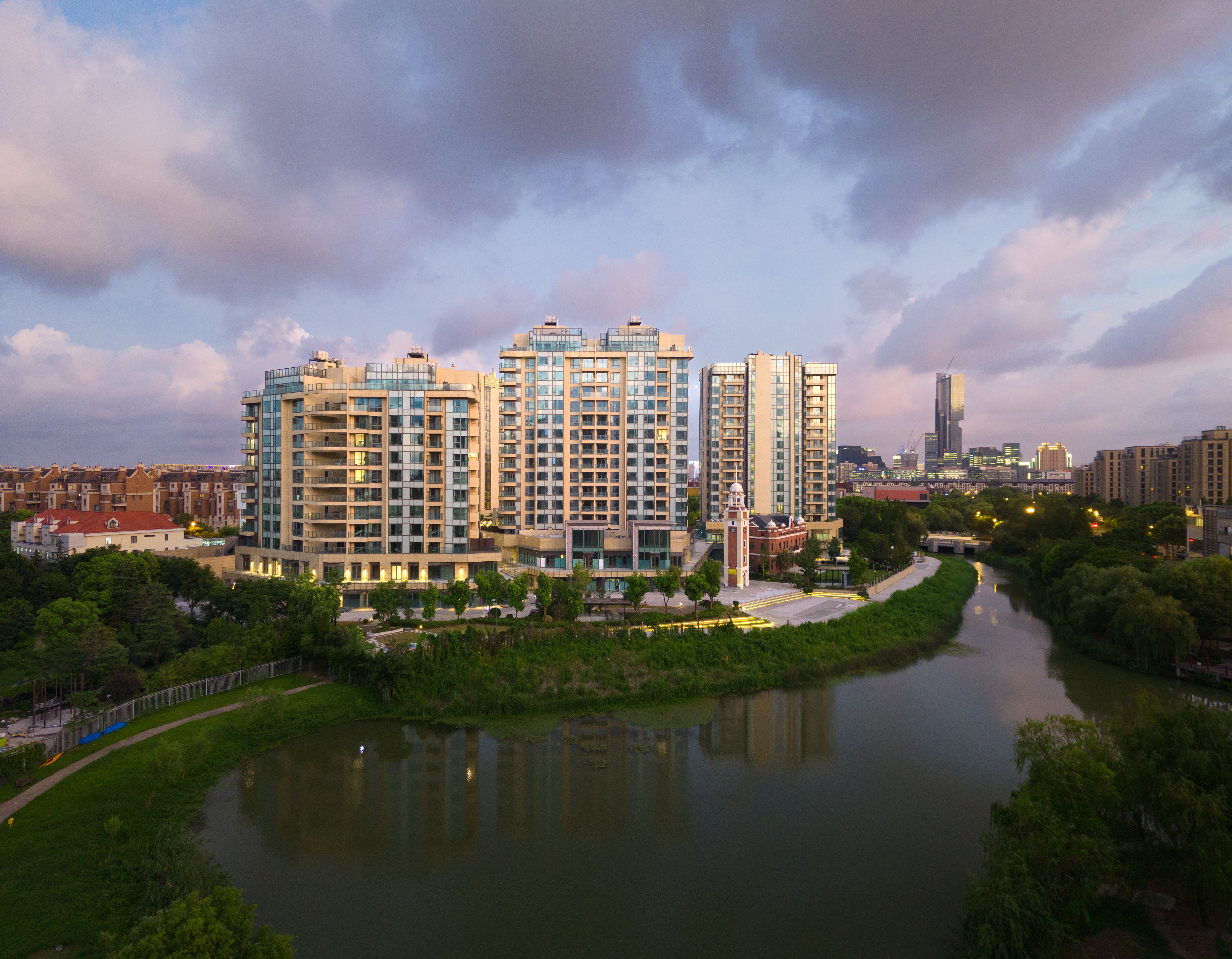 Aerial view of modern mid-rise residential buildings situated along a calm riverbank during sunset. The buildings are surrounded by lush greenery and city infrastructure, with a partly cloudy sky casting a warm glow over the scene. A city skyline is visible in the background.
