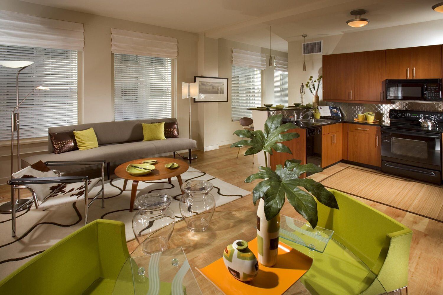 A kitchen that is next to a living room with green and grey couches along with wooden tables.