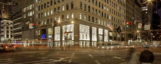 View of a corner building in a busy city at night. Cars drive past in the foreground.