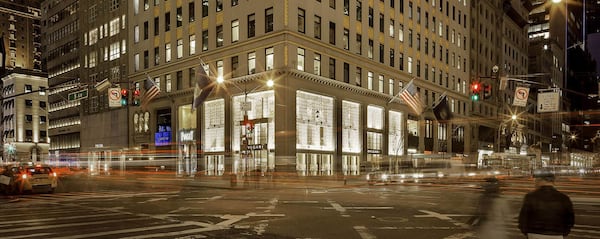 View of a corner building in a busy city at night. Cars drive past in the foreground.