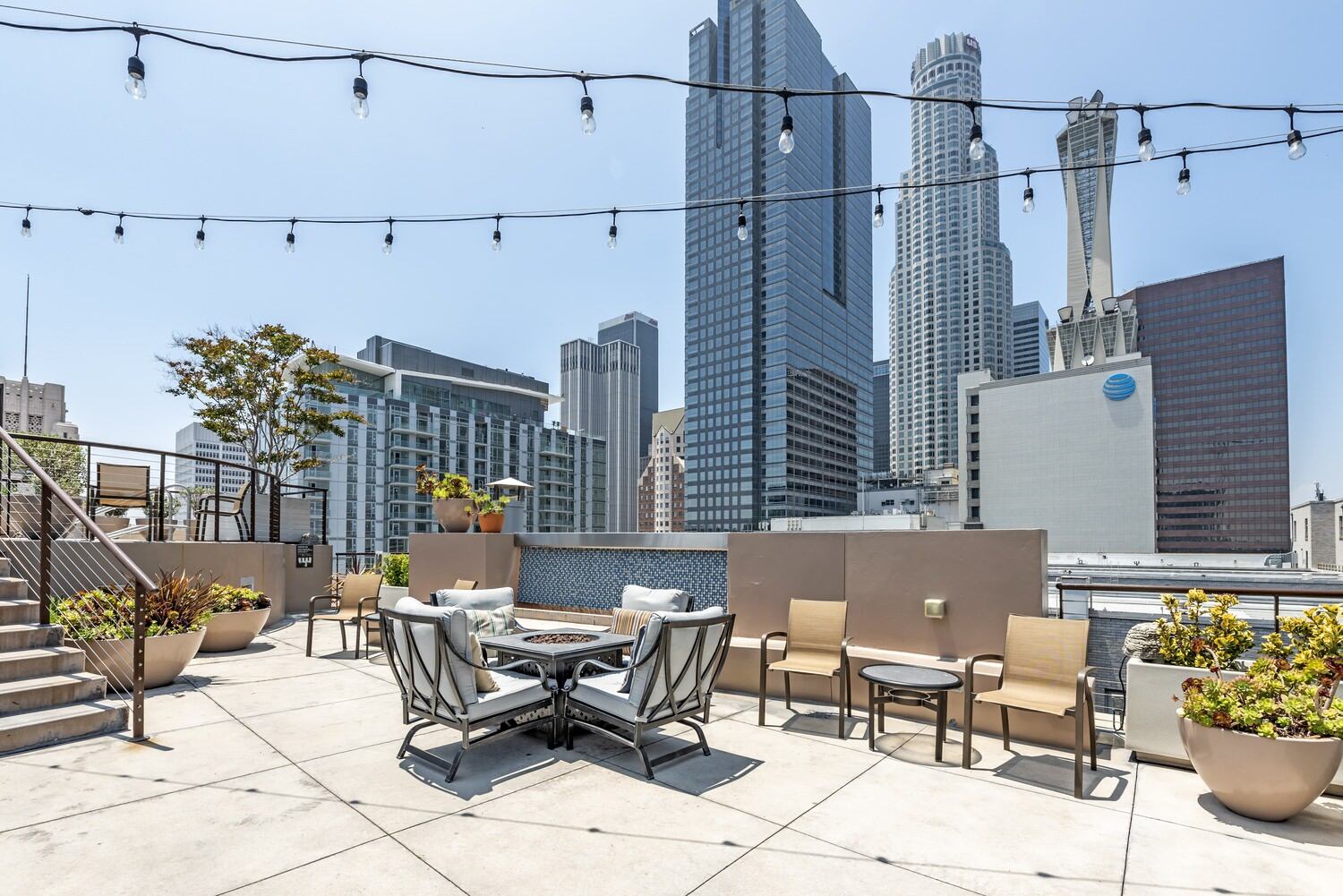 A rooftop patio that has tables and chairs as well as a fire pit, there are also planters with flowers in them.